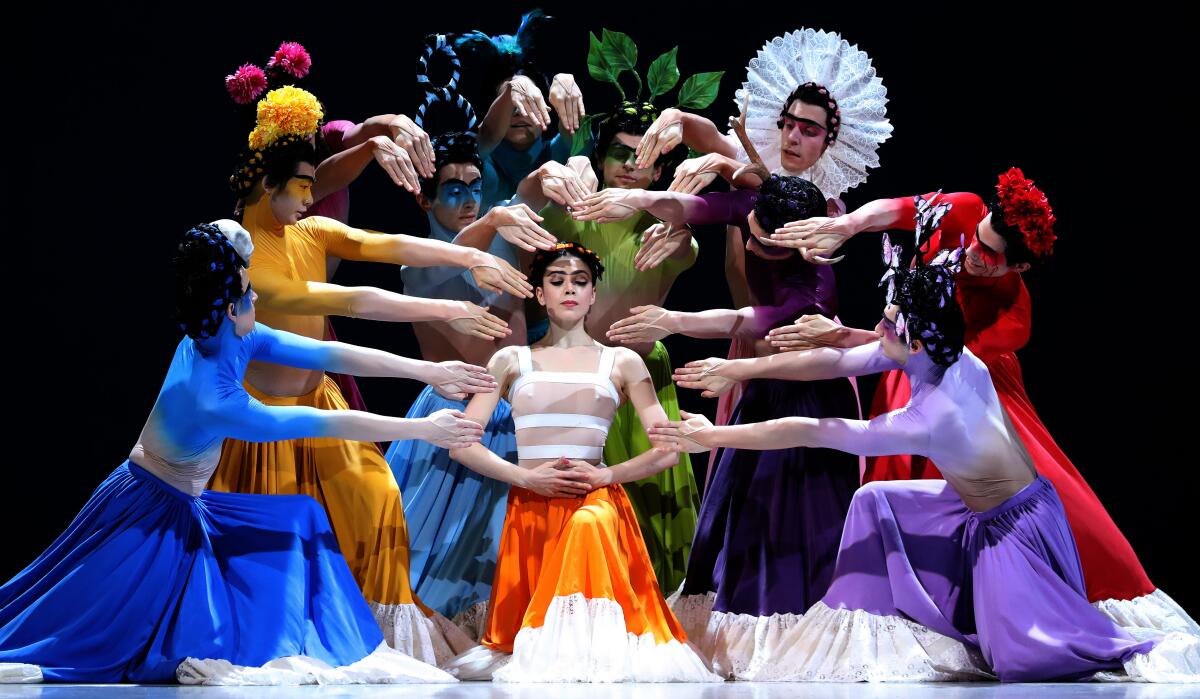 A woman is surrounded by dancers in bright dresses with outstretched hands.