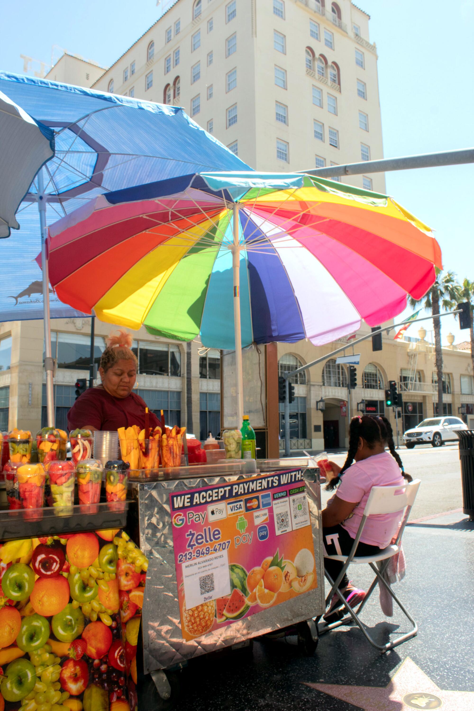 A vendor sits with her stand, which features a rainbow-colored umbrella, along a city street.