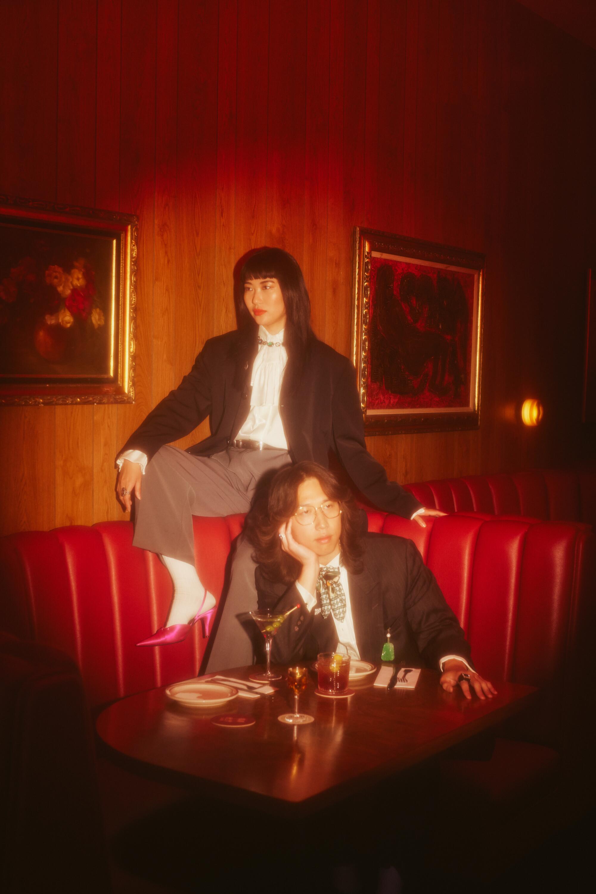 An Asian couple sit at restaurant booth with drinks, each looking off to the side. They are wearing vintage suits.