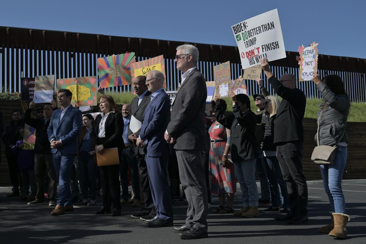 People hold protest signs near a border wall.