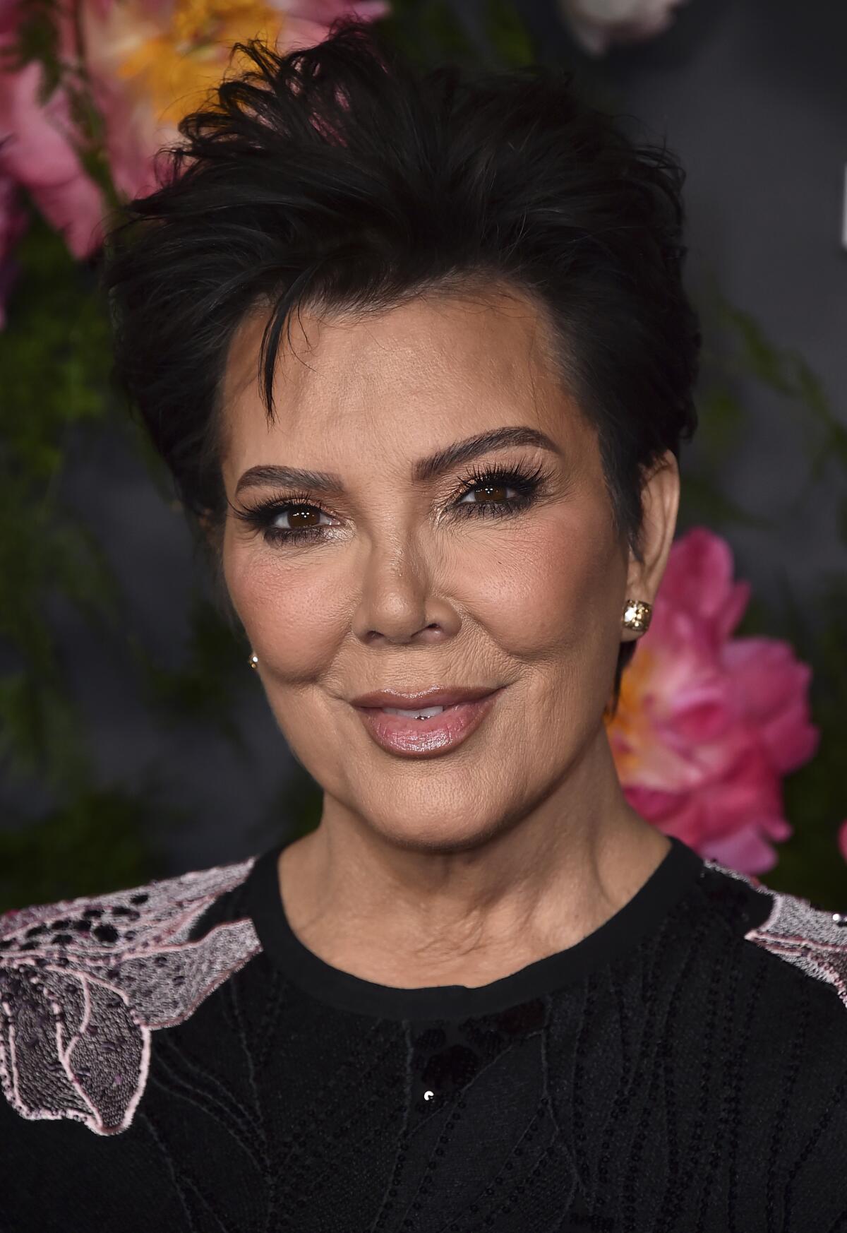 Kris Jenner smiles while wearing a black dress with white floral embroidery 