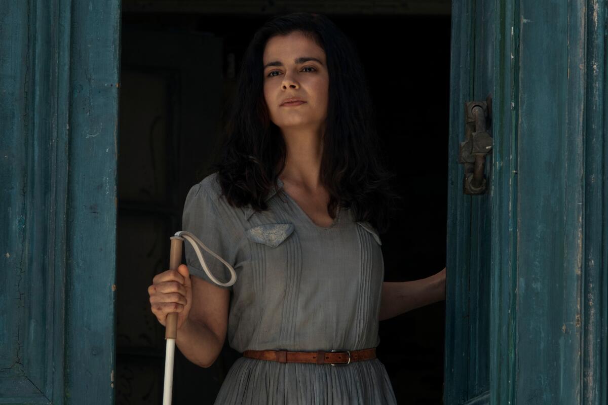 A young woman with dark hair and a light blue dress stands in a doorway holding a cane.