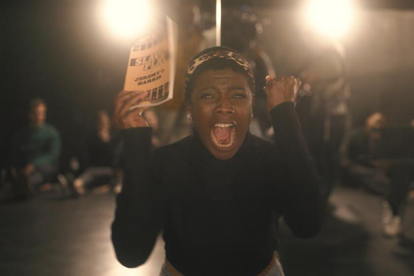 A Black woman holding a script and facing the camera appears to scream in rage as two hot spotlights glow from from behind. 