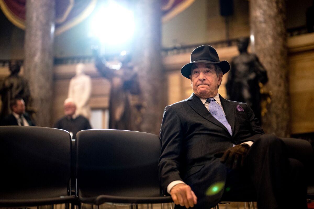 Paul Pelosi, wearing a suit and a hat sits on a row of chairs inside a tall room with stone columns