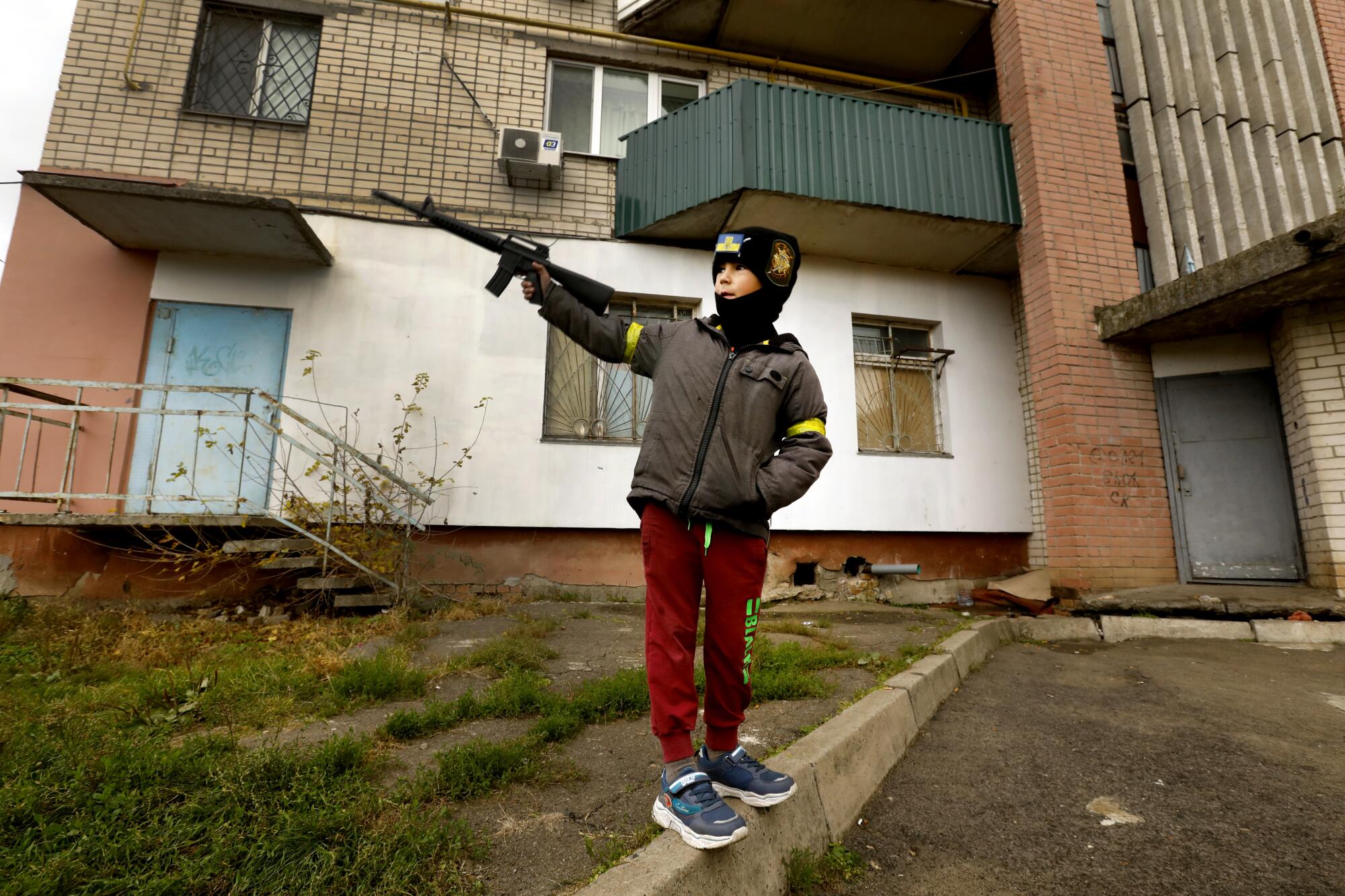 A boy in a dark cap, brown jacket and red pants brandishes a toy gun in front of a building