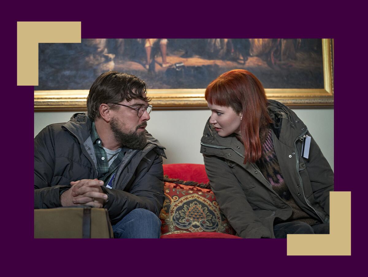 A man and a woman in winter clothing sit on a couch looking at each other with alarmed expressions.