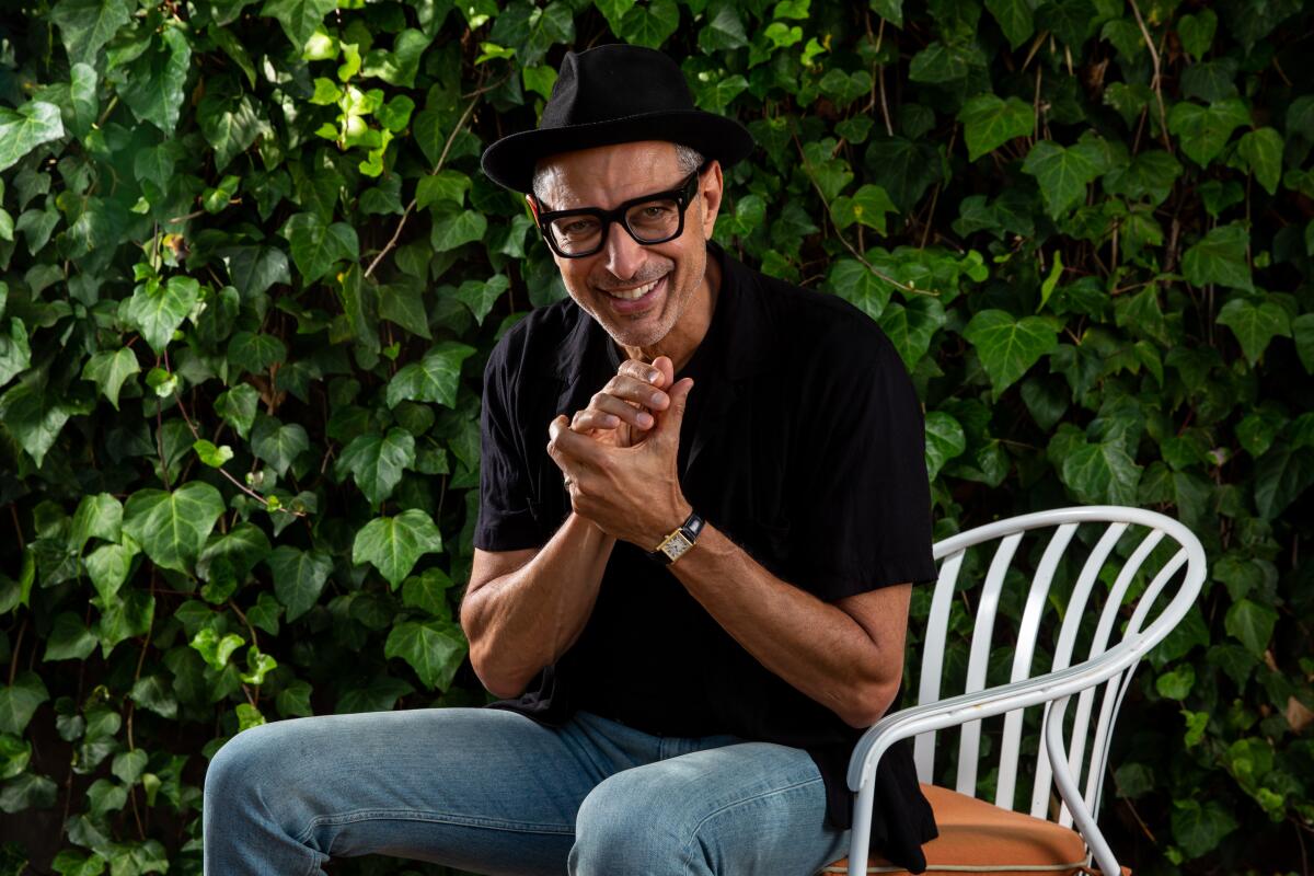 Jeff Goldblum, wearing glasses, sits outdoors in a chair, clasping his hands in glee.