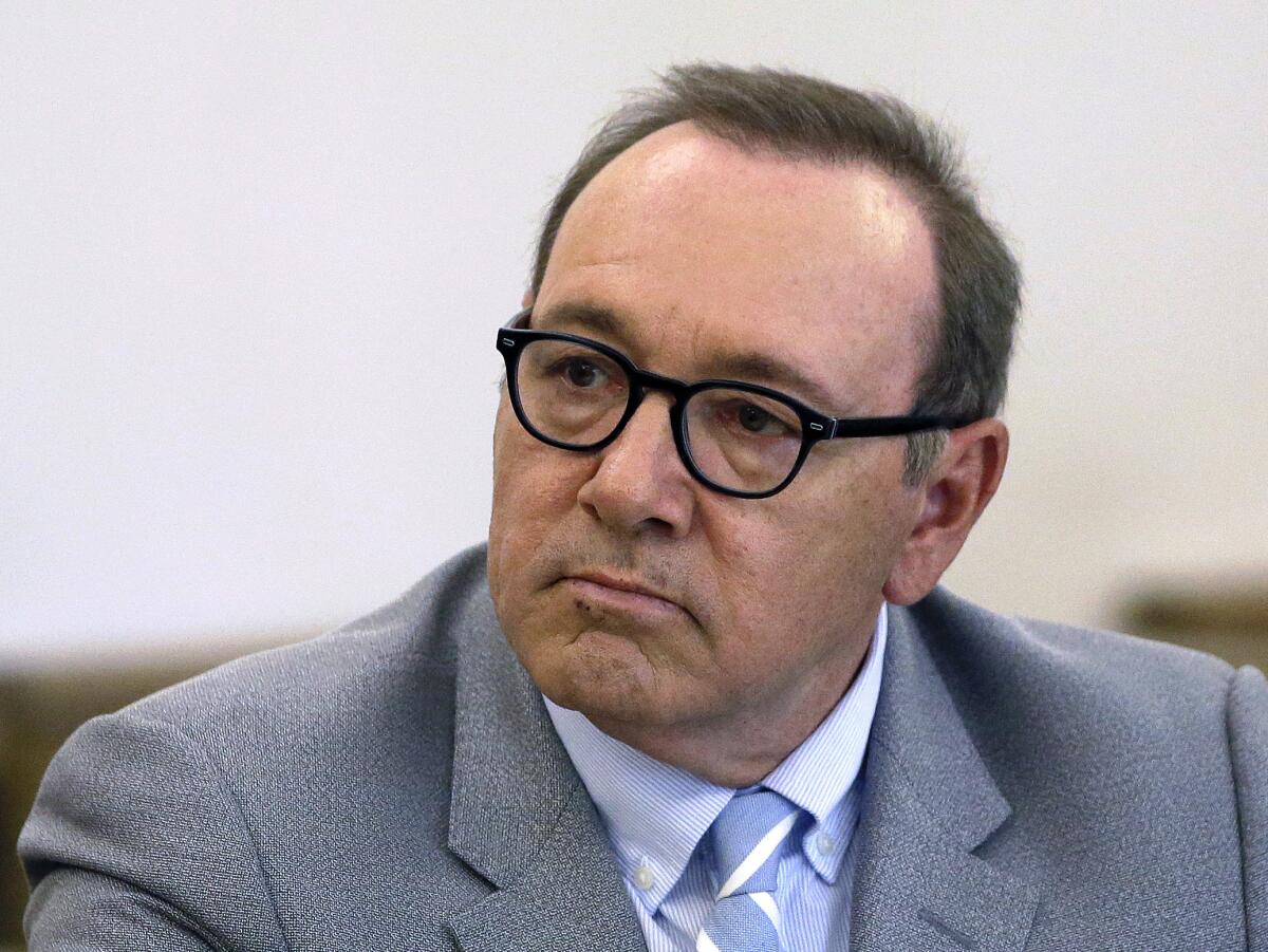 A man in dark-rimmed eyeglasses and a gray suit frowns in court