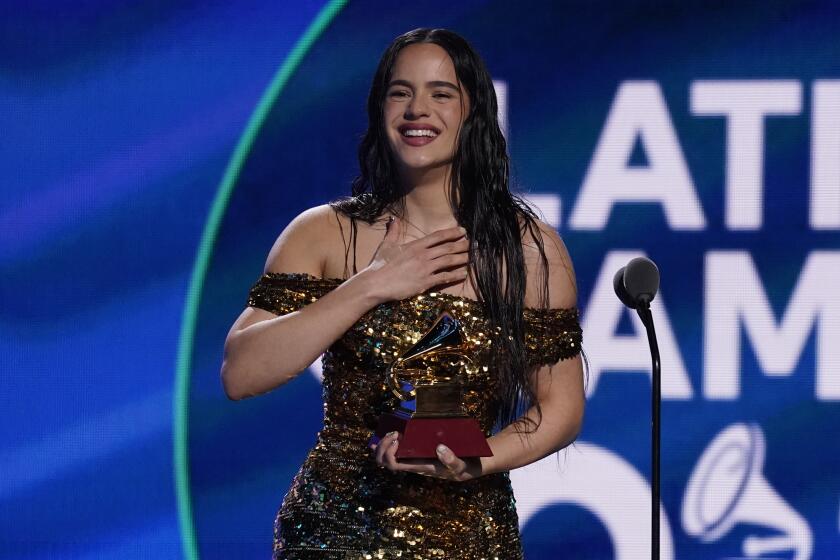 A woman with long brown hair wearing a gold dress, smiling and holding an award in front of a microphone