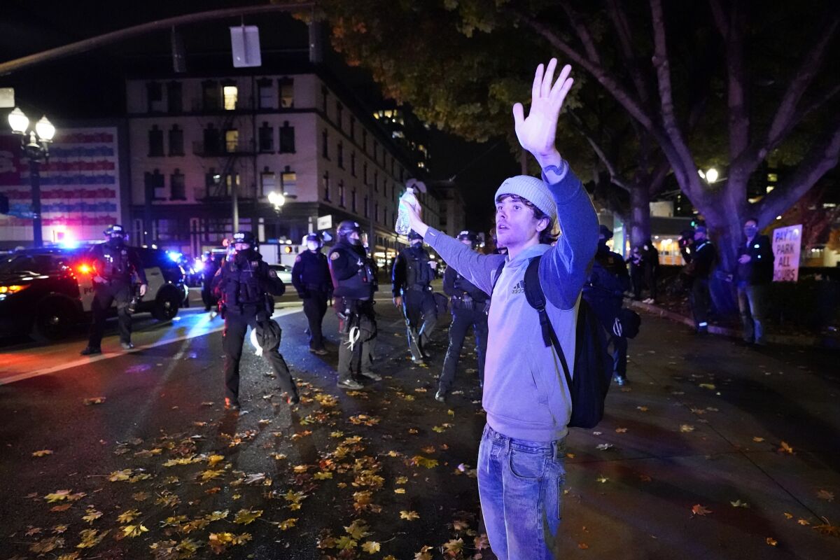 A man holding a water bottle raises his arms in front of a group of police wearing riot gear