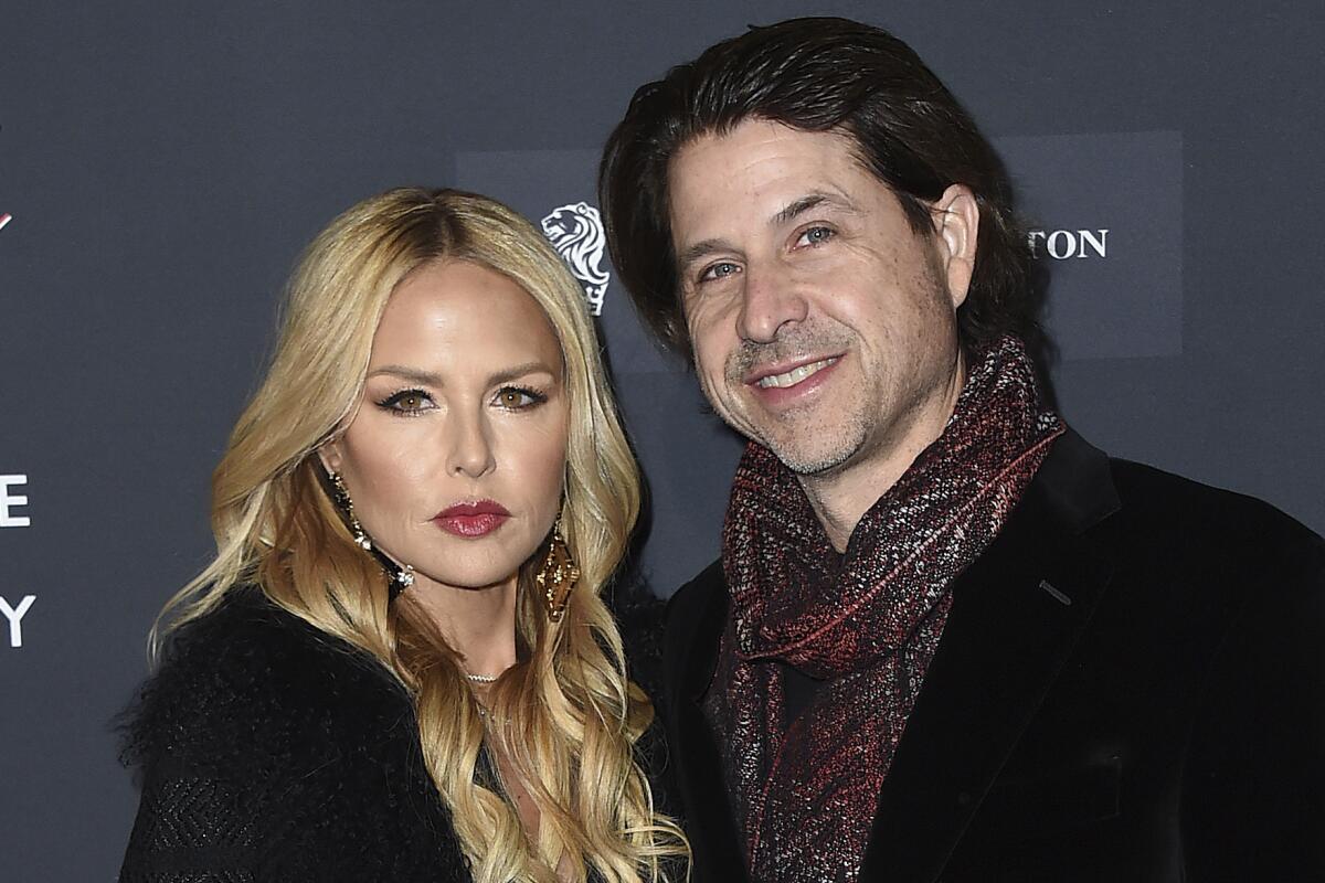 Rachel Zoe looking serious in a black coat next to a smiling Rodger Berman in a black coat and patterned scarf