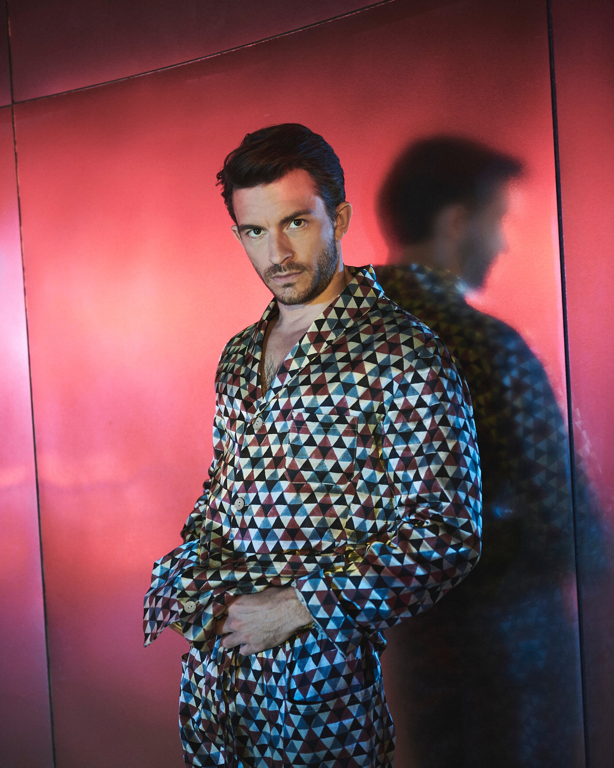 A man in patterned silk clothing leans up against a red, reflective wall.