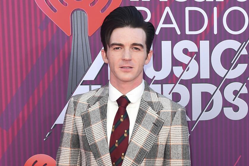 Drake Bell in a gray and beige plaid suit with a red tie posing against a purple and red backdrop with white text