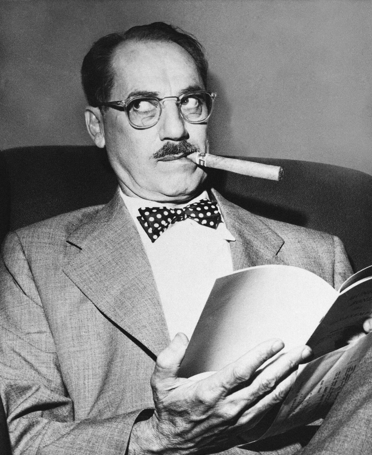 A man with a moustache, glasses and a cigar sits with a pamphlet in his hands and looks to the side.  
