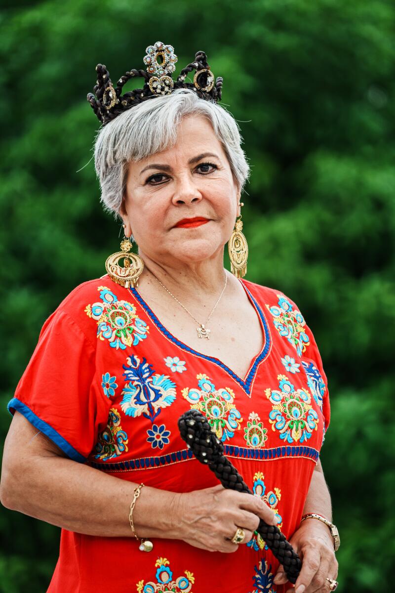 A gray-haired woman in a red dress wearing a crown and holding a wand, both made from woven vanilla pods.