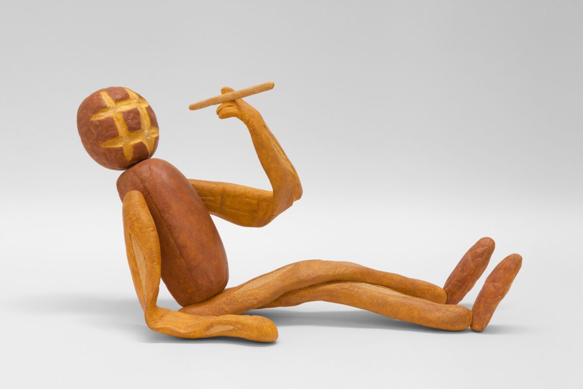 A reclining humanoid figure, holding aloft a cigar, is carved to look as if it is made of loaves of bread.