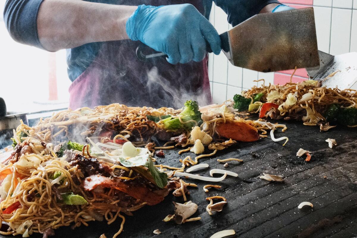 Two gloved hands hold spatulas over noodles and vegetables on a large flat circular plancha at Three Flames in Westchester.