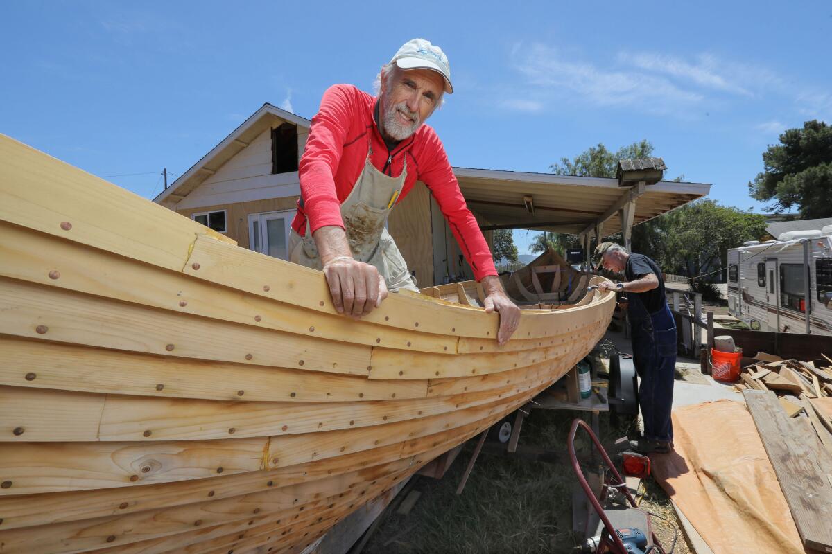 A man smiles over the side of a boat being built in a backyard.