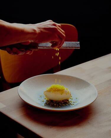 A hand shaves a cured egg yolk over a dish of panna cotta.