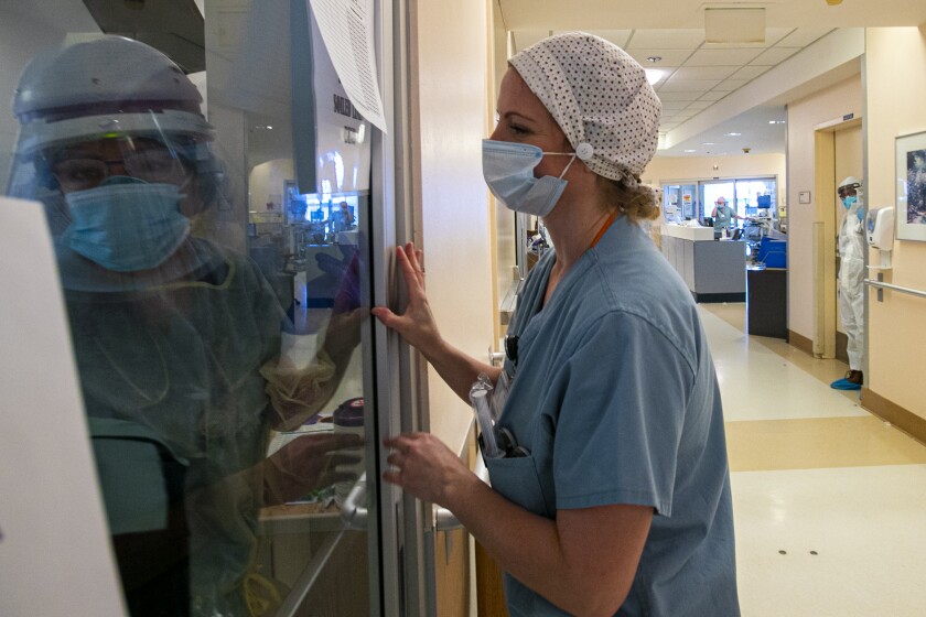 A nurse looks through a window at another health worker in full protective gear.