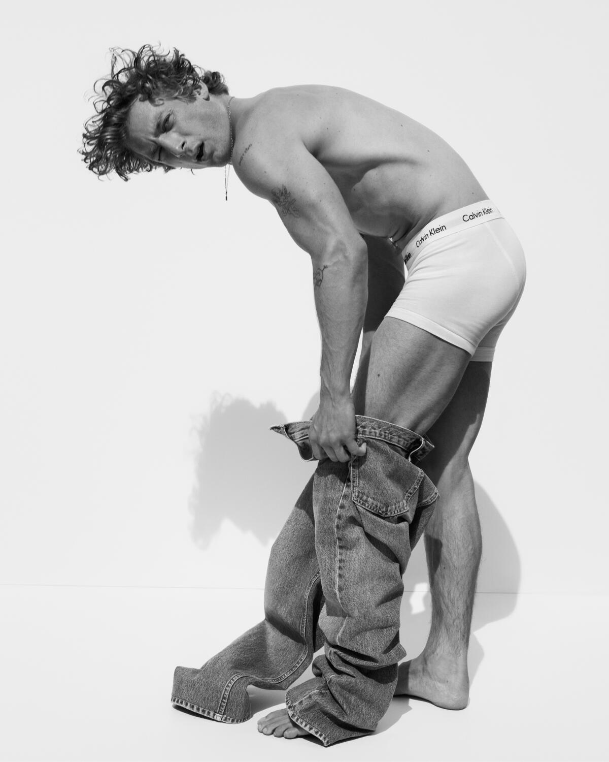 Jeremy Allen White stepping into a pair of blue jeans with no shirt on. 