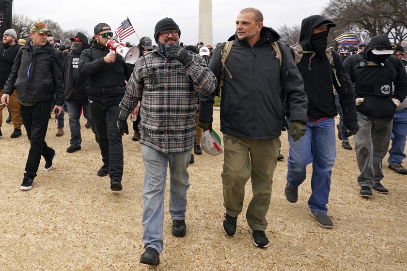 A group of men wearing black and carrying U.S. flags walk on dry grass with the Washington Monument in the background