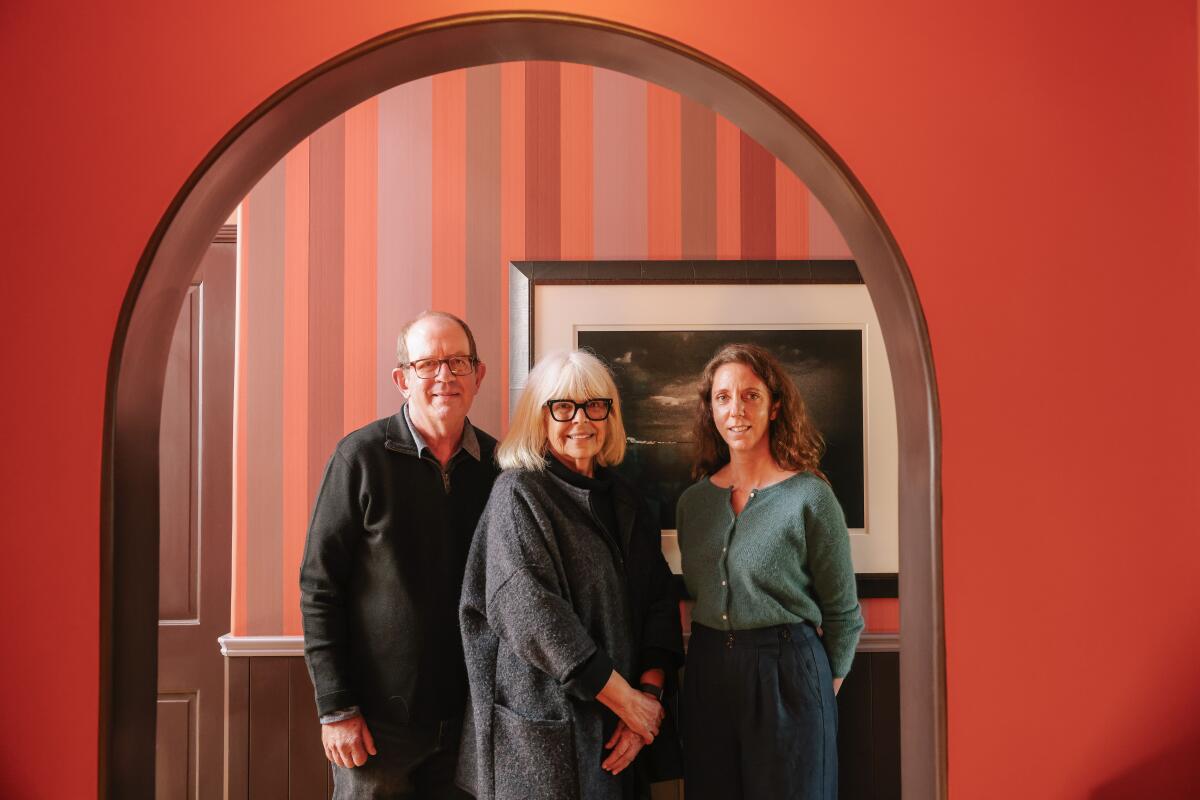 Three people pose under an archway in a home with reddish walls and a striped wall behind them.