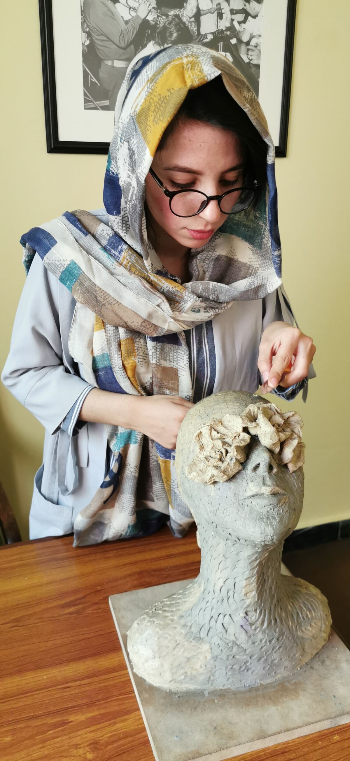 A woman wearing glasses and head scarf looks down at a bust of a person with covered eyes 