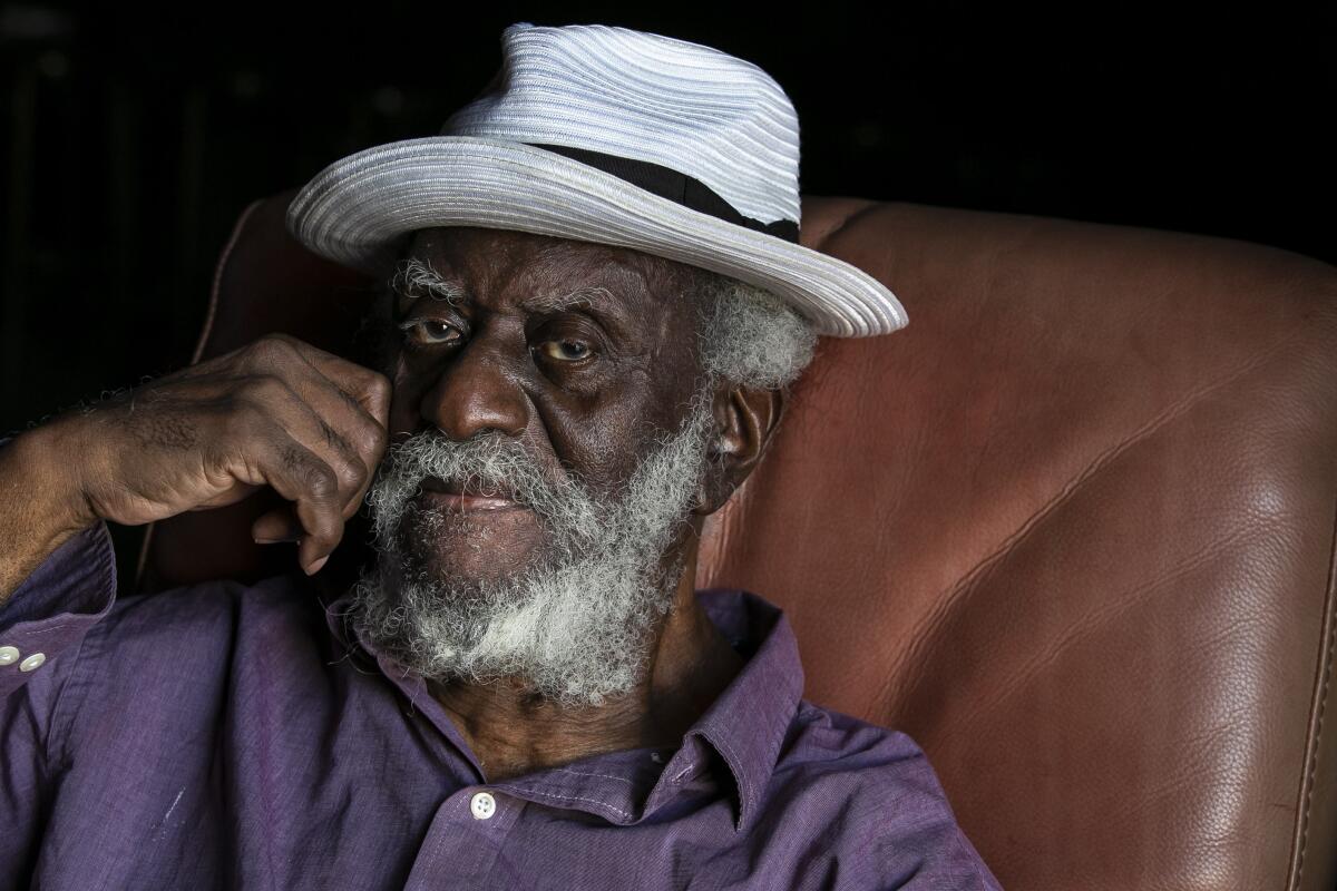 A Black man with a white beard and hat sits on a couch, looking at the camera. 