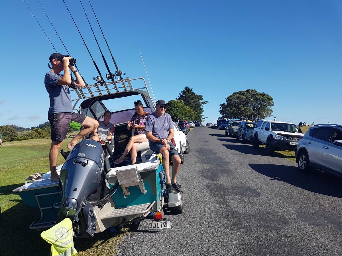 A man looks through binoculars while standing in a boat with other people on a road with parked cars 