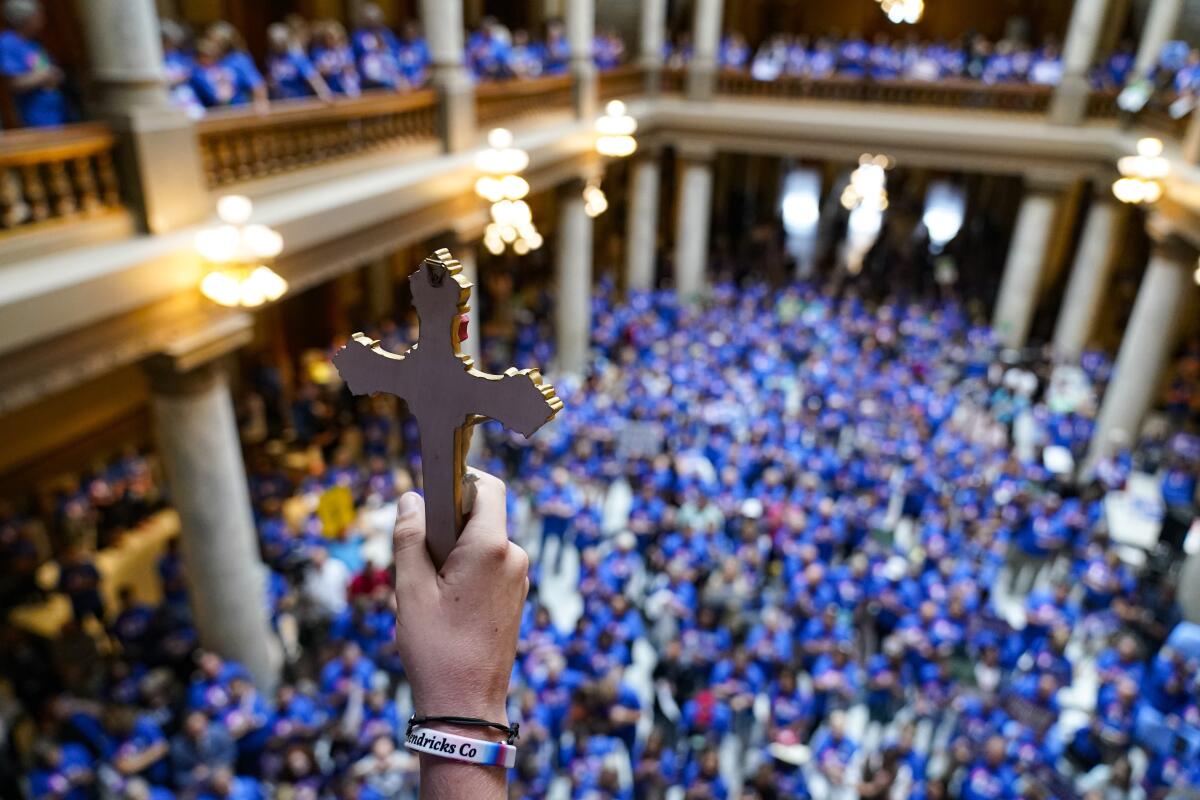 A hand holds a crucifix from a balcony above a sea of blue-clad ralliers.