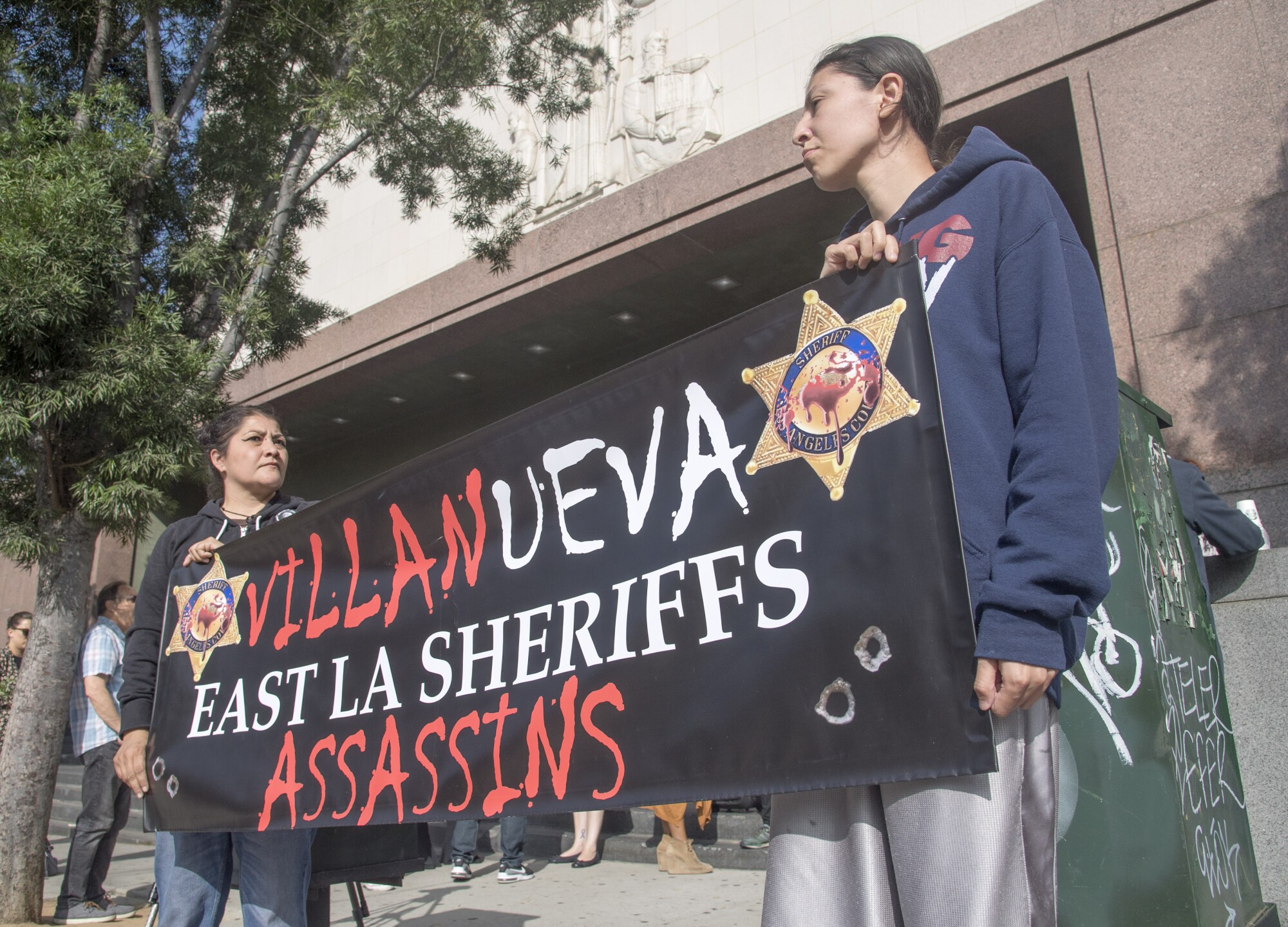 Valerie Vargas and Stephanie Luna hold a sign with the words "Villanueva East LA Sheriffs Assassins" on it.