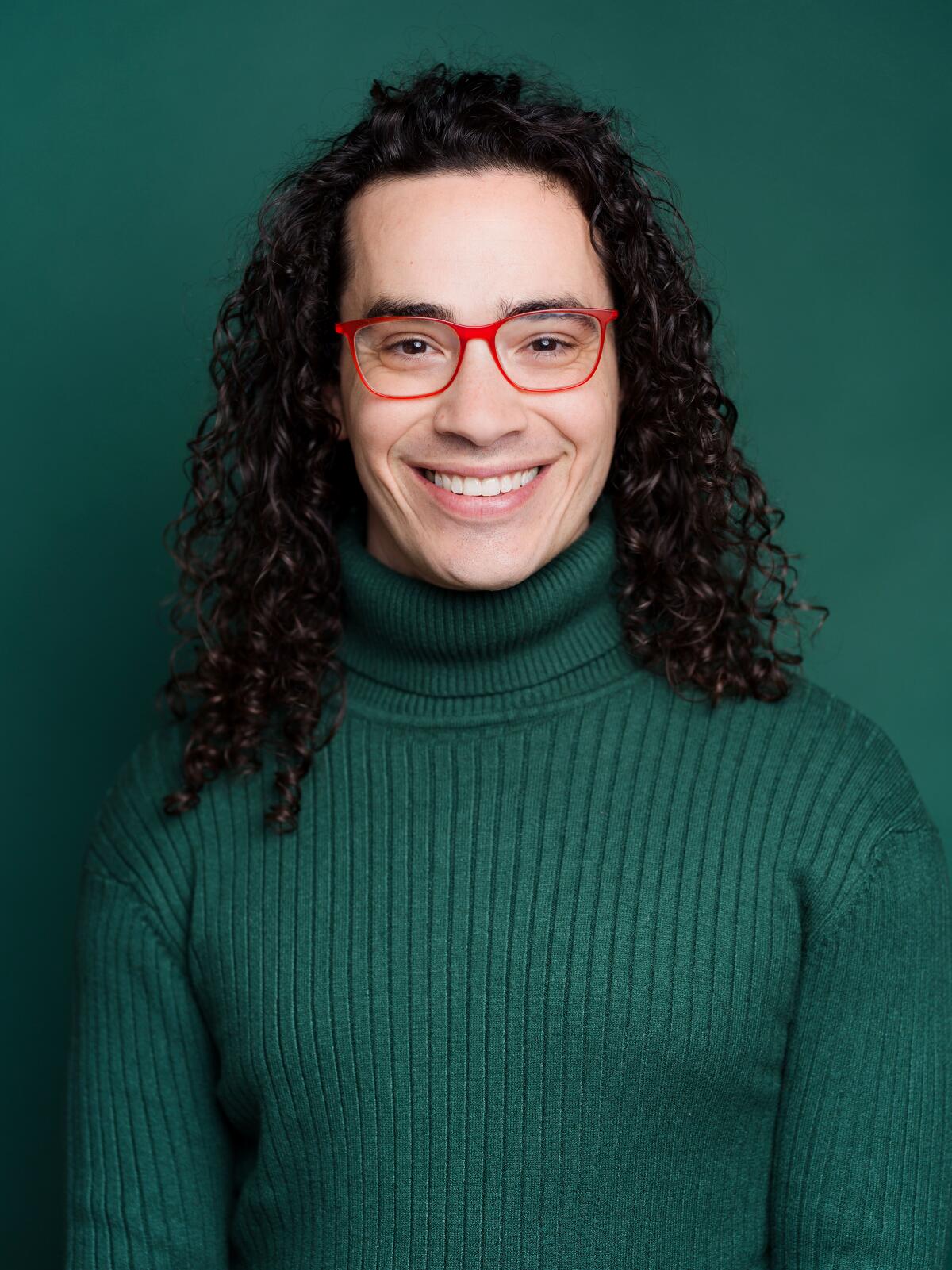 Jake Brasch, in a green turtleneck sweater and red-framed glasses, smiles at the camera. 