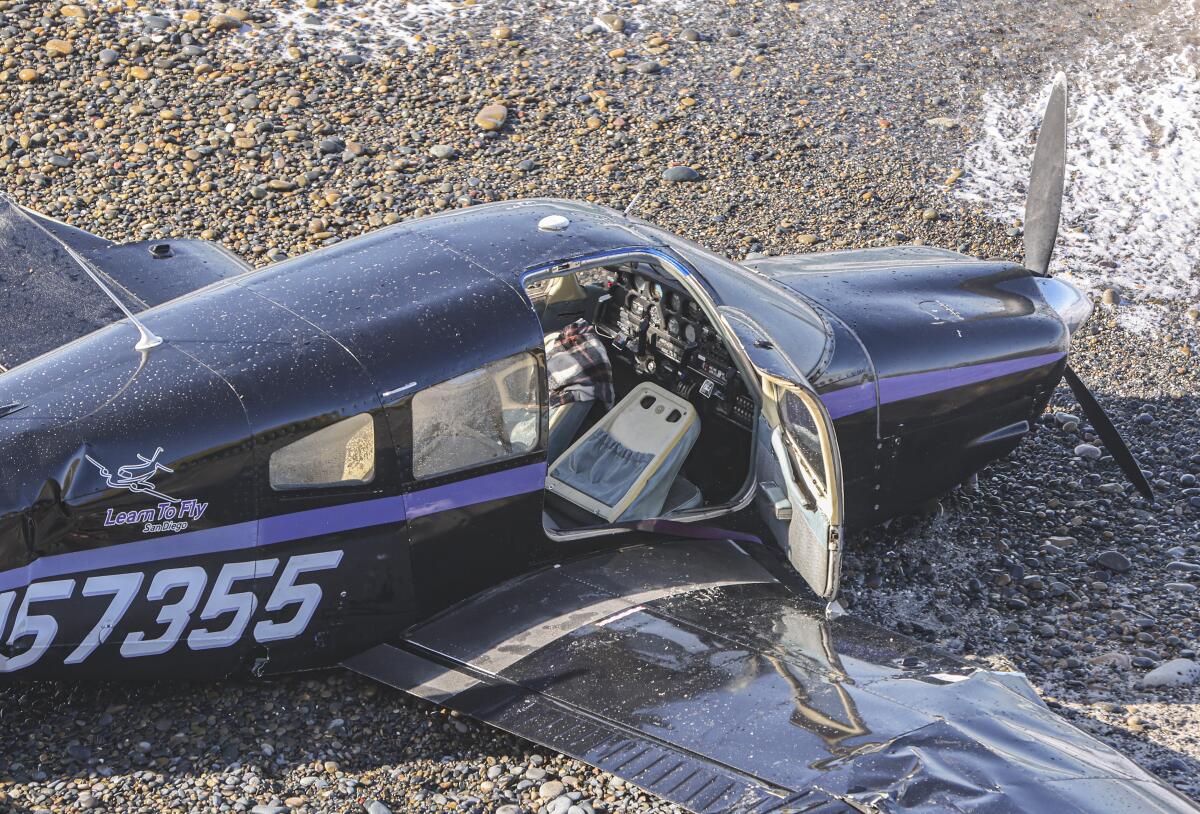 A crashed small plane lies on the beach with its open door showing the seats and instruments inside