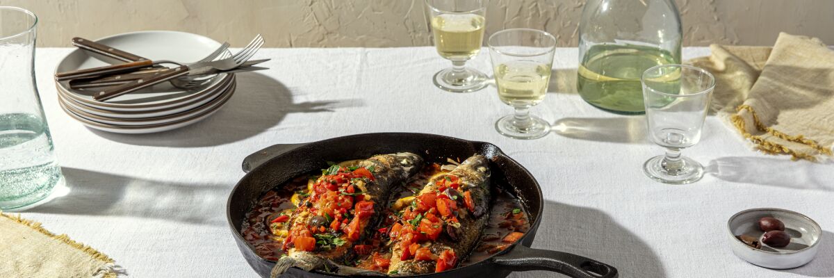  Roasted Branzino with Tomatoes and Olives in a cast-iron pan on a table set with wine glasses and a stack of plates.