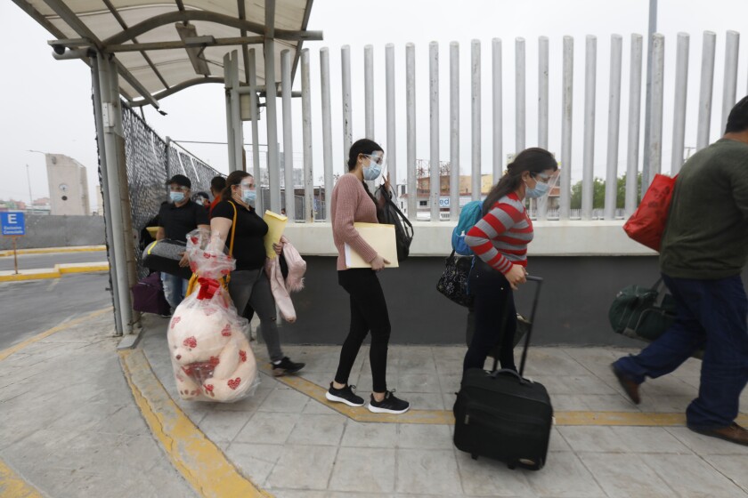 People wearing masks and carrying belonging walk by a fence