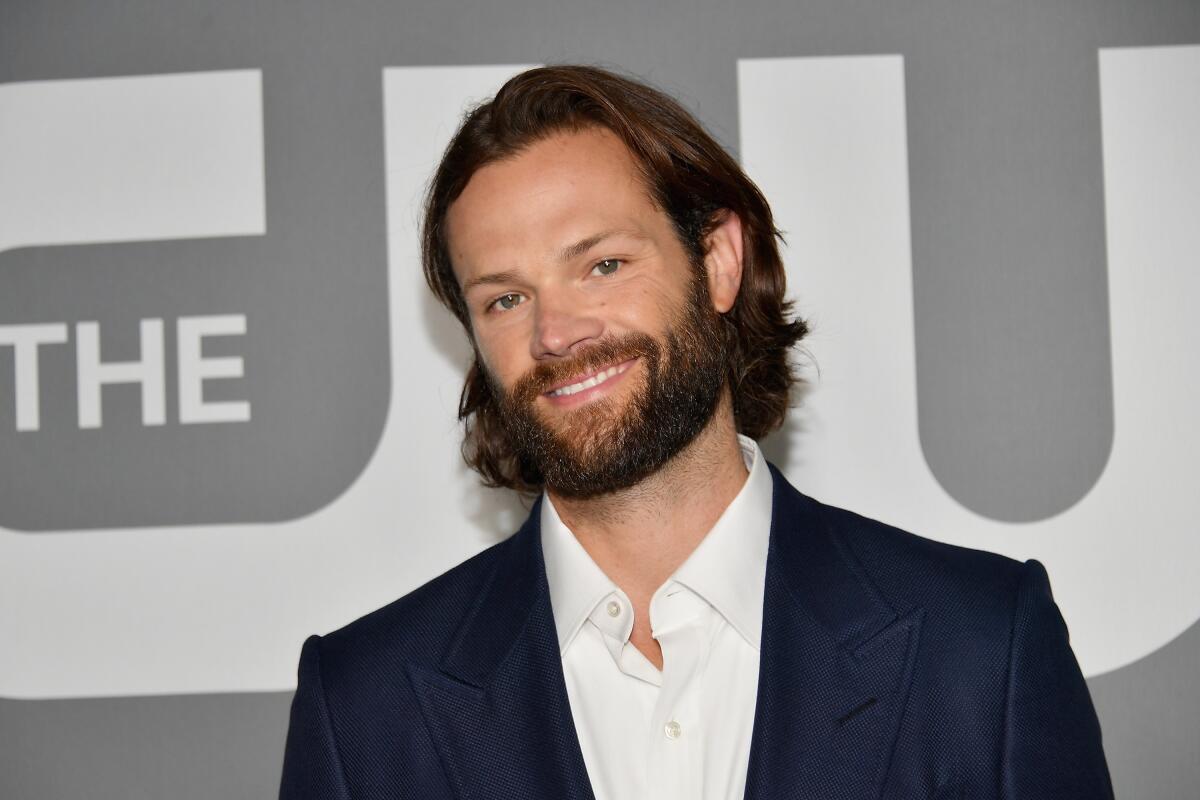Jared Padalecki smiling with long hair and a beard while clad in a suit jacket and a white button down with no tie