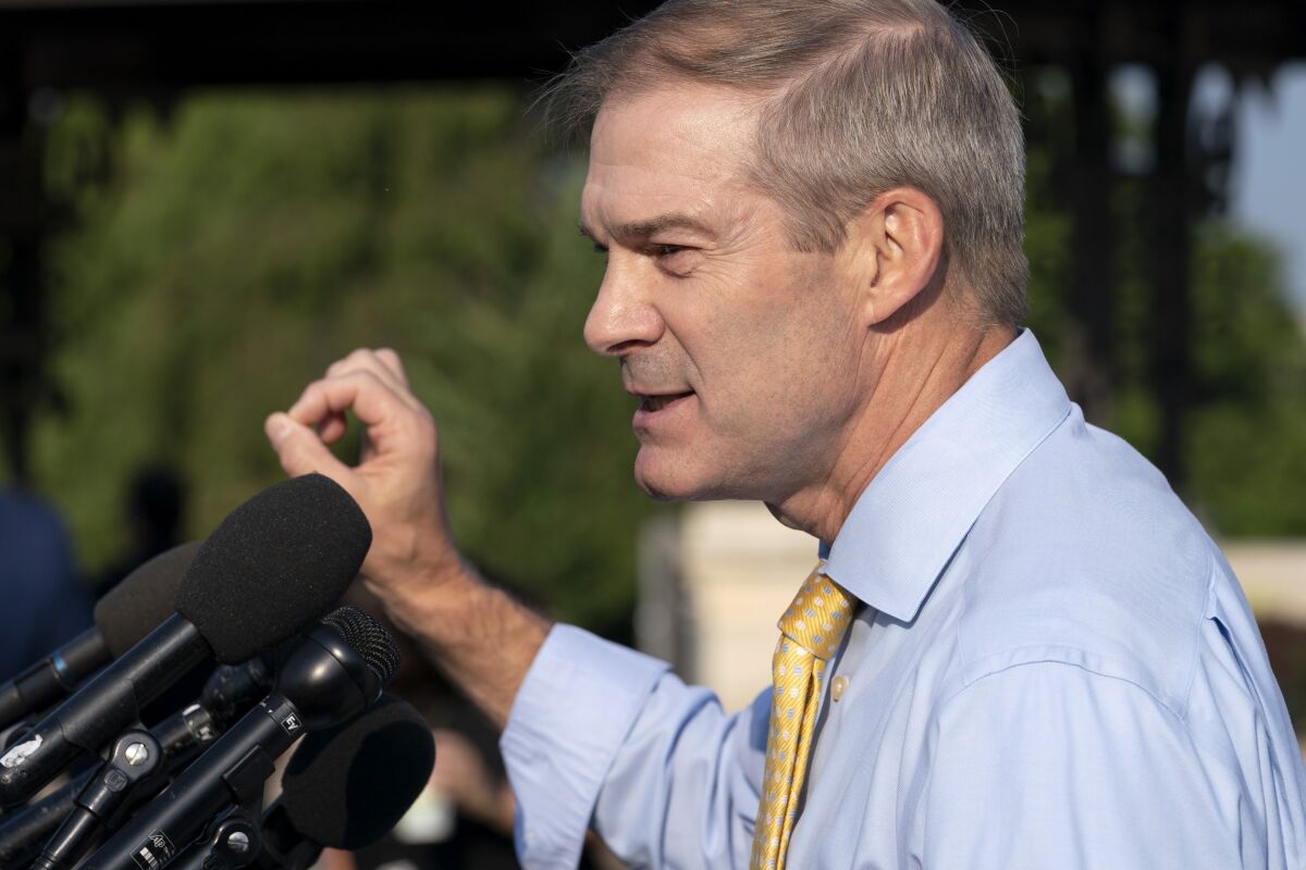 A side view of a man in a light blue shirt and yellow tie speaking before microphones 