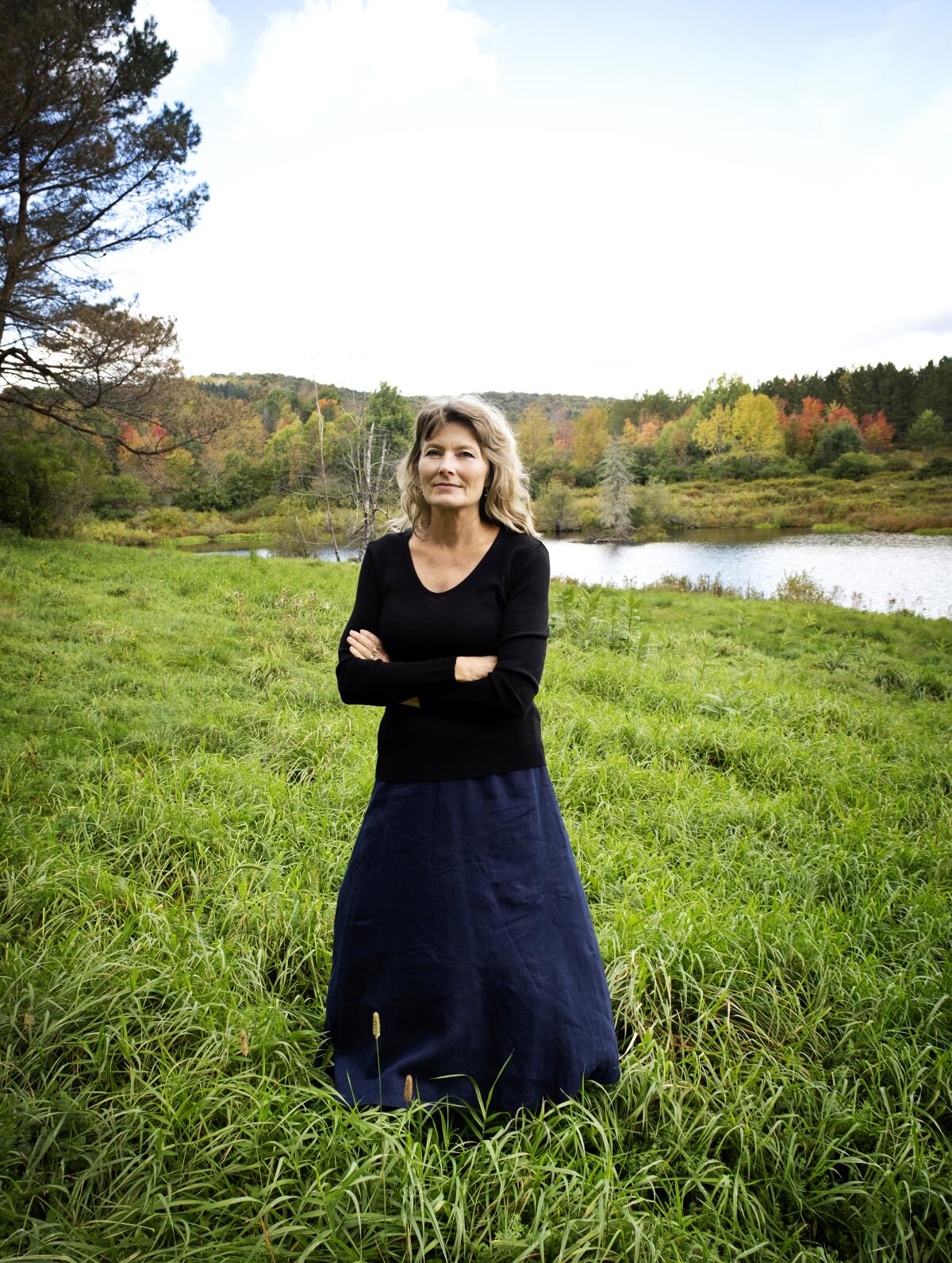 A woman in a dark-colored shirt and skirt stands in a field with water in the background.
