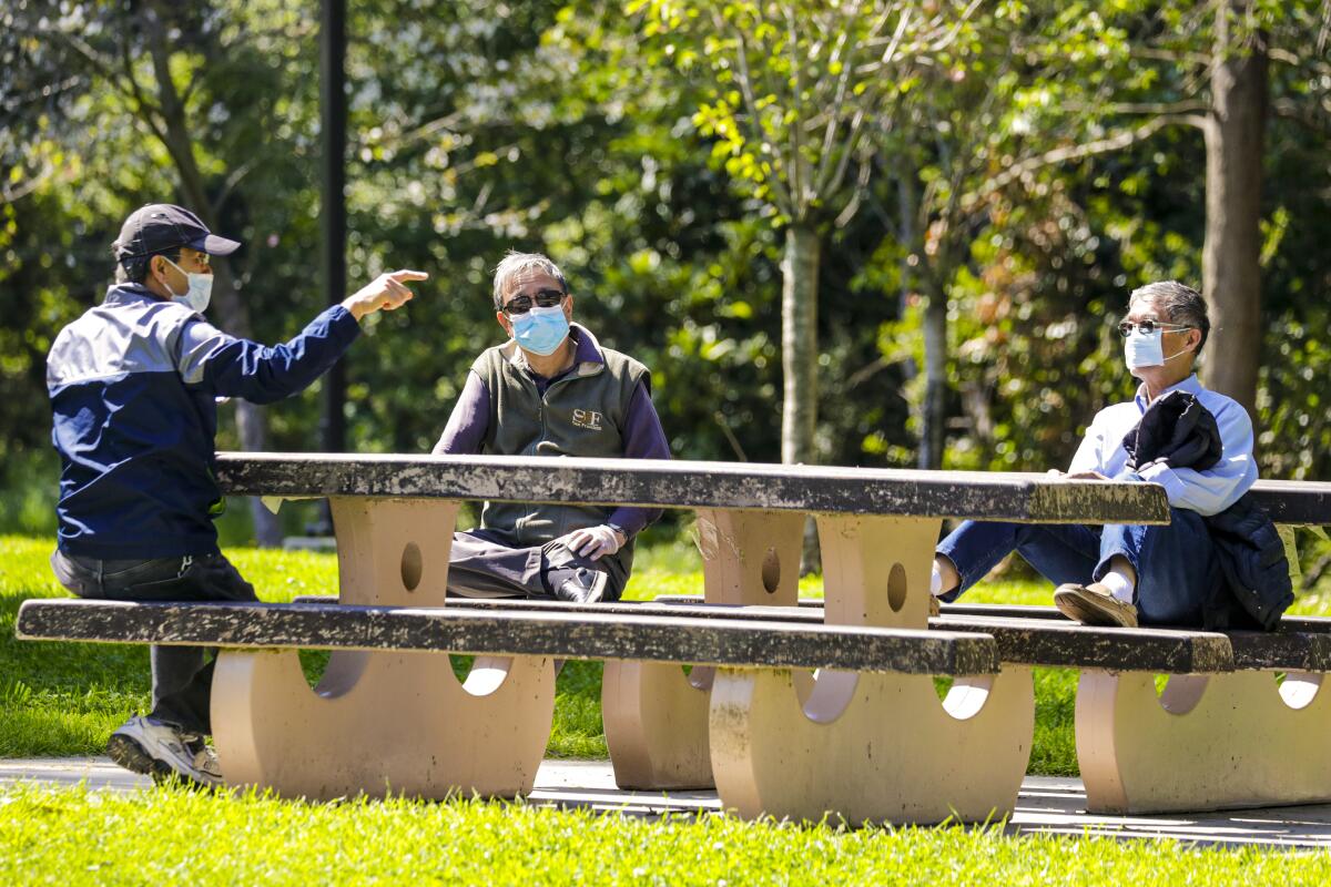 Three friends covering their faces with masks and sitting at safe distance from each other chat sitting at a picnic table.