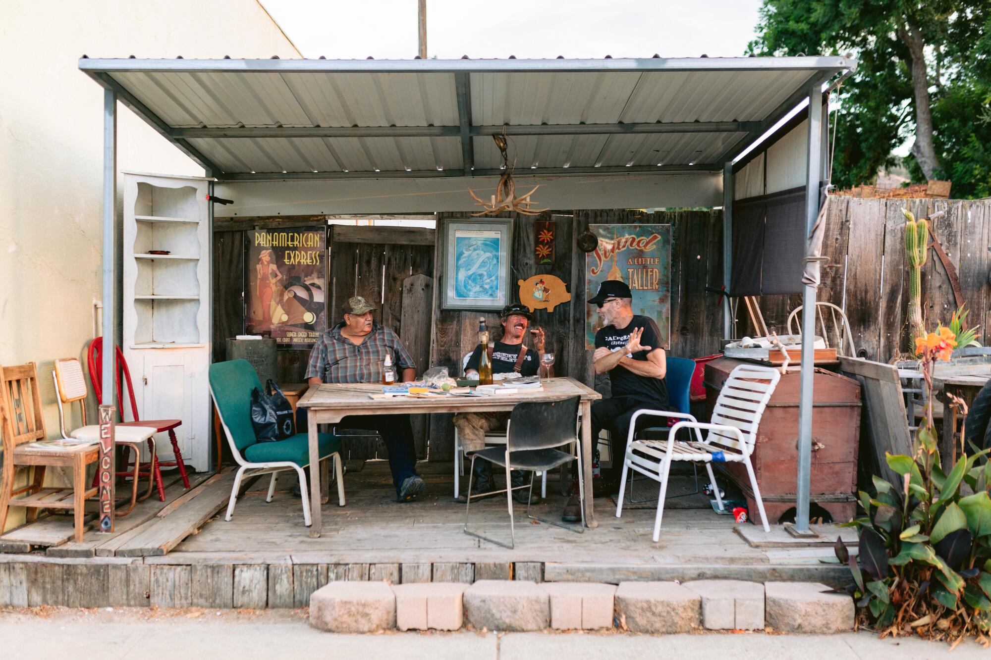 Three men sit at a sheltered table off a sidewalk in a small town.