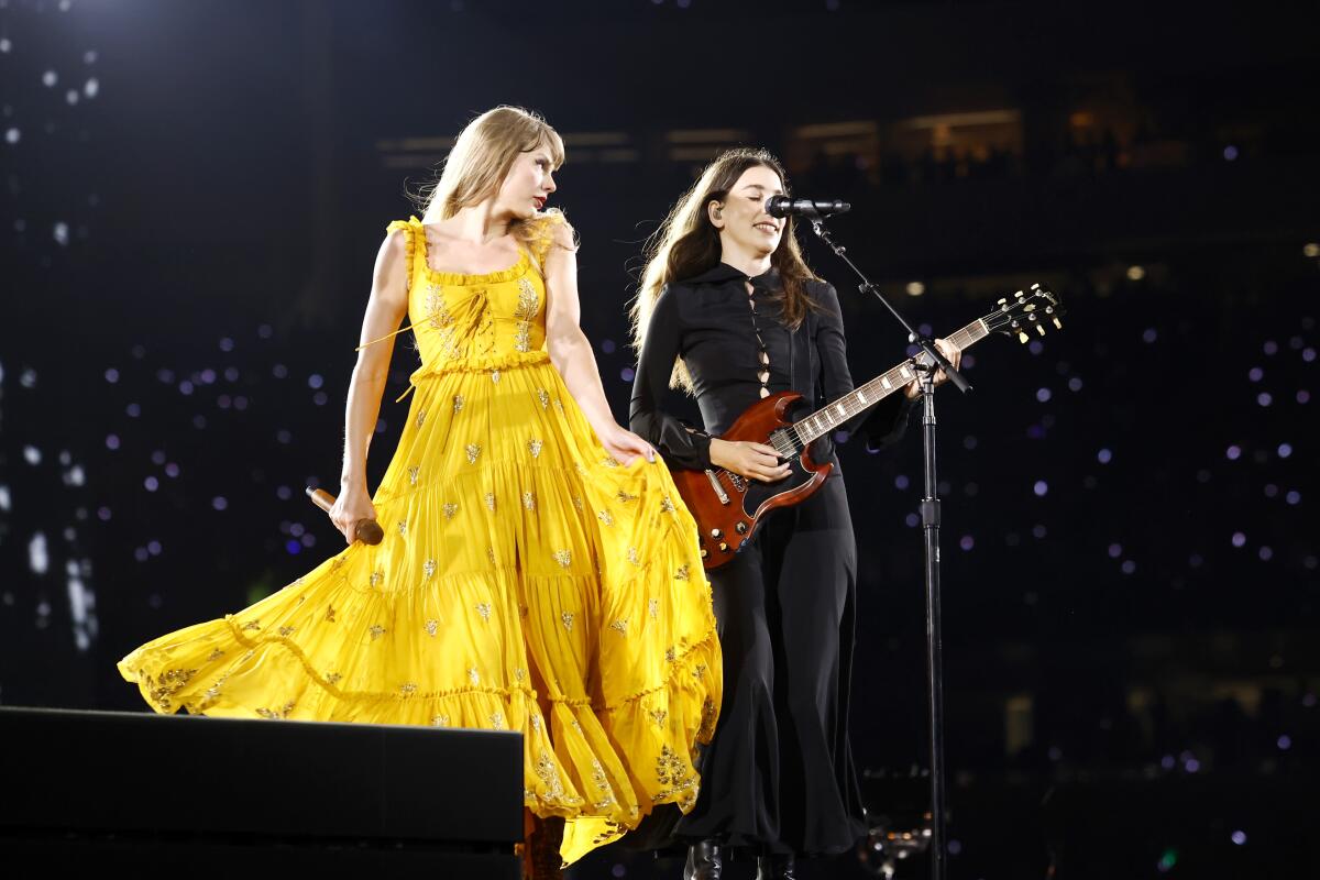 Taylor Swift in a floor length yellow dress and Danielle Haim on guitar perform onstage.