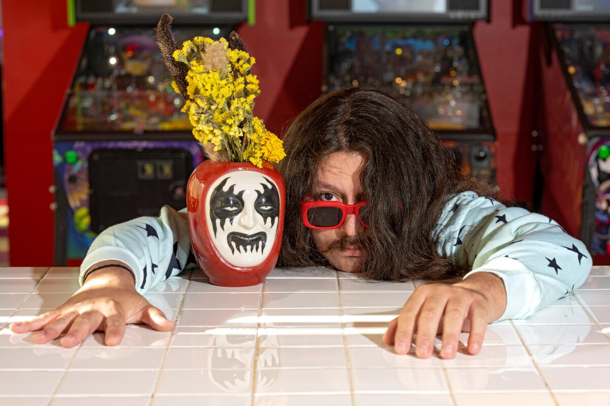 A long-haired man poses with a vase painted with a creepy face and holding yellow flowers.