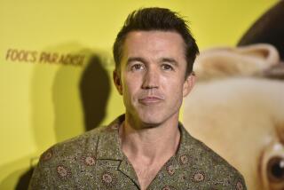 Rob McElhenney poses in a patterned, collared shirt against a yellow background.