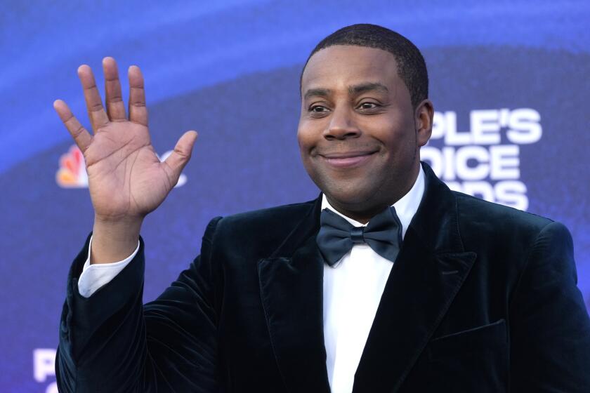 Keenan Thompson is waving and smiling with his mouth closed while wearing a black tuxedo with a blue bowtie.