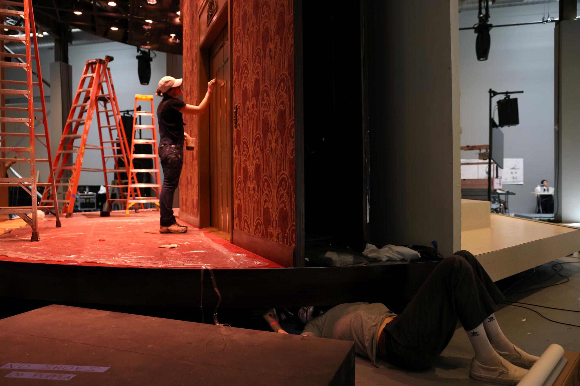 A woman in a cap paints a stage door while another works under the stage. 