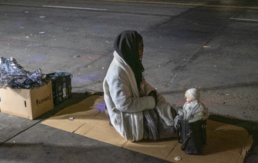 A woman sits next to a doll perched on a purse on the sidewalk on April 22, 2021.