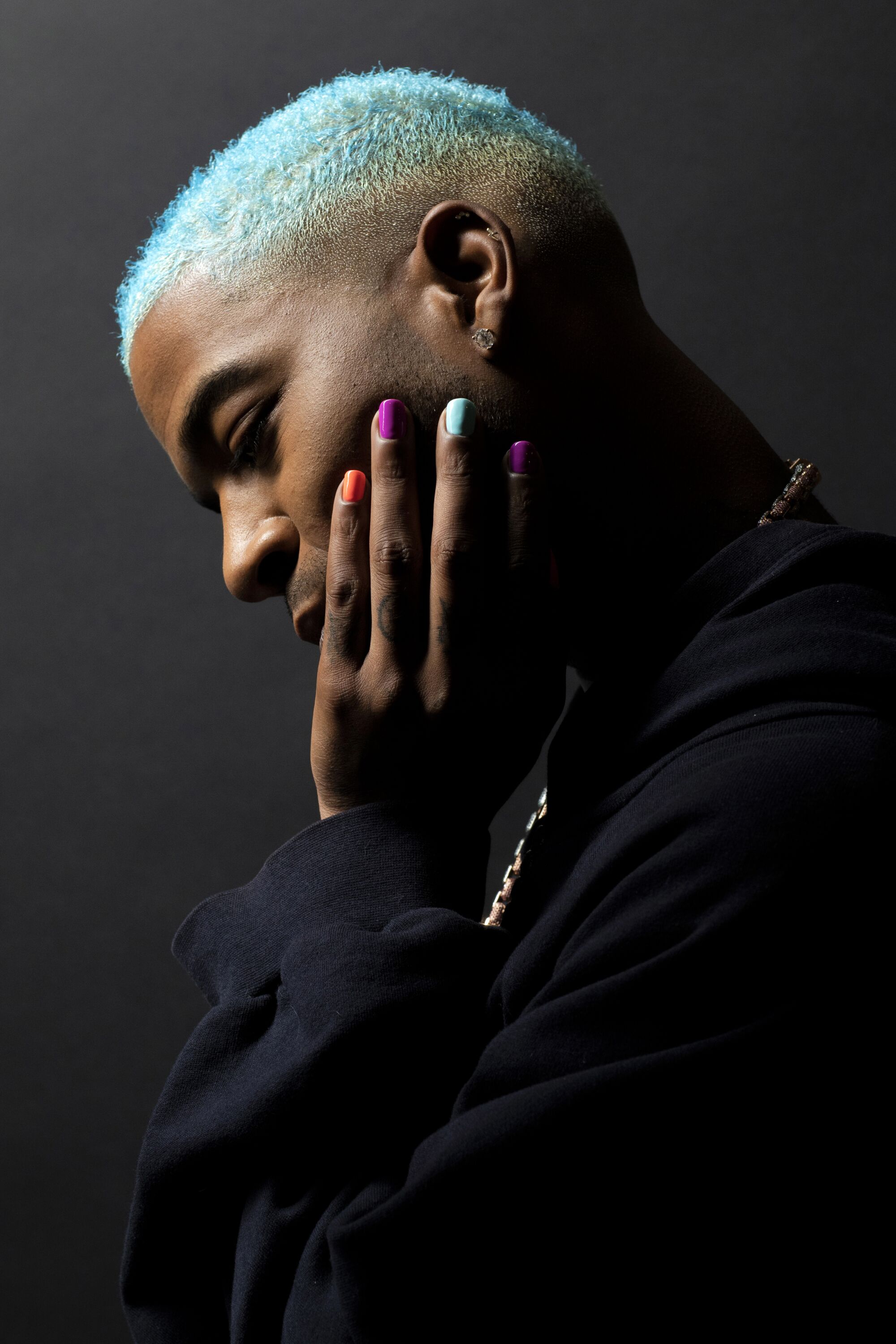 A man with blue hair and painted nails poses for a portrait on a dark backdrop.