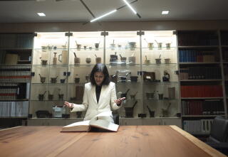 A woman in a cream-colored jacket looks down on an open book on a table, with a glass medical display case behind her