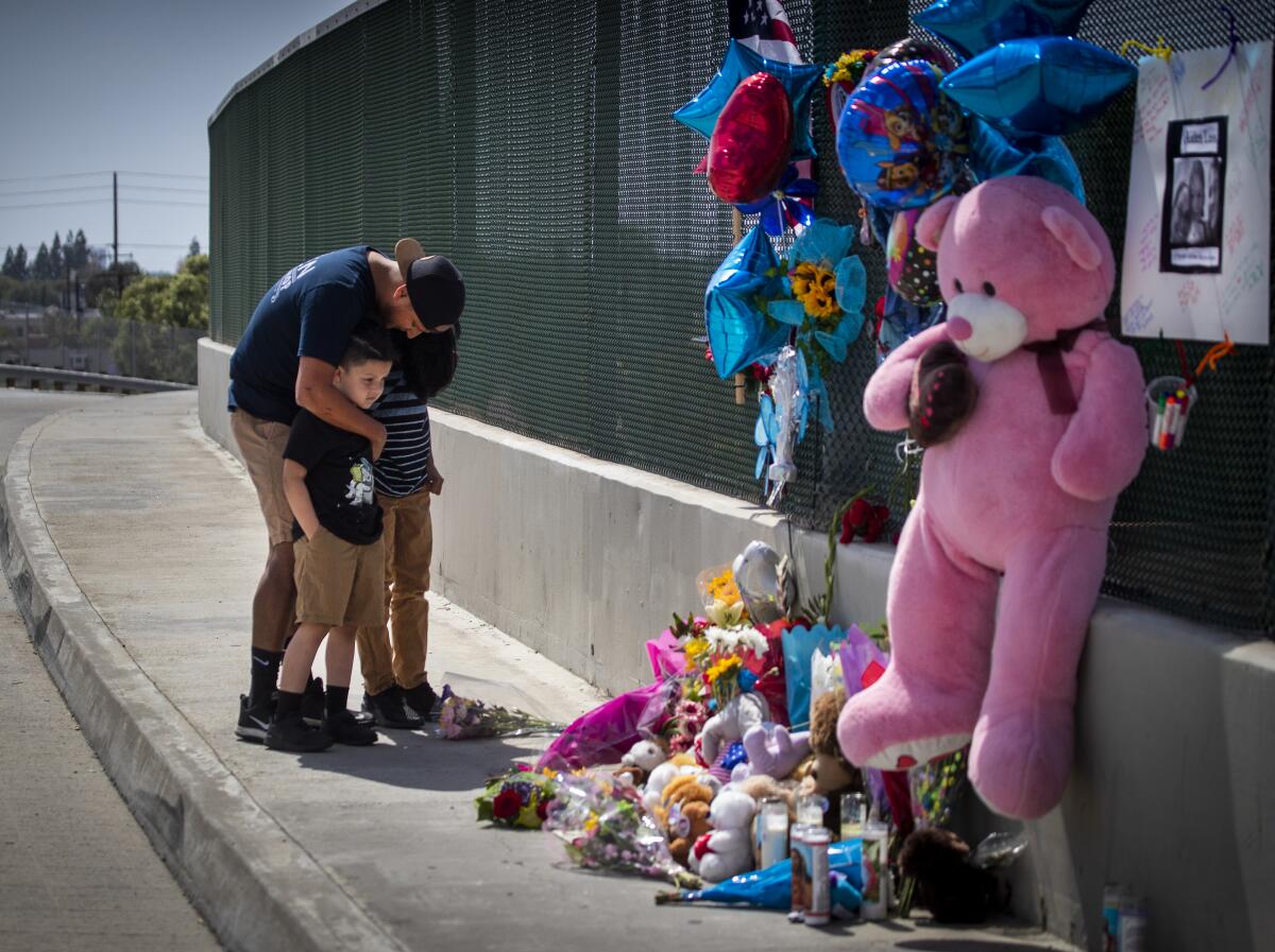 A man hugs two boys next to a fence with a memorial made up of balloons, candles and teddy bears